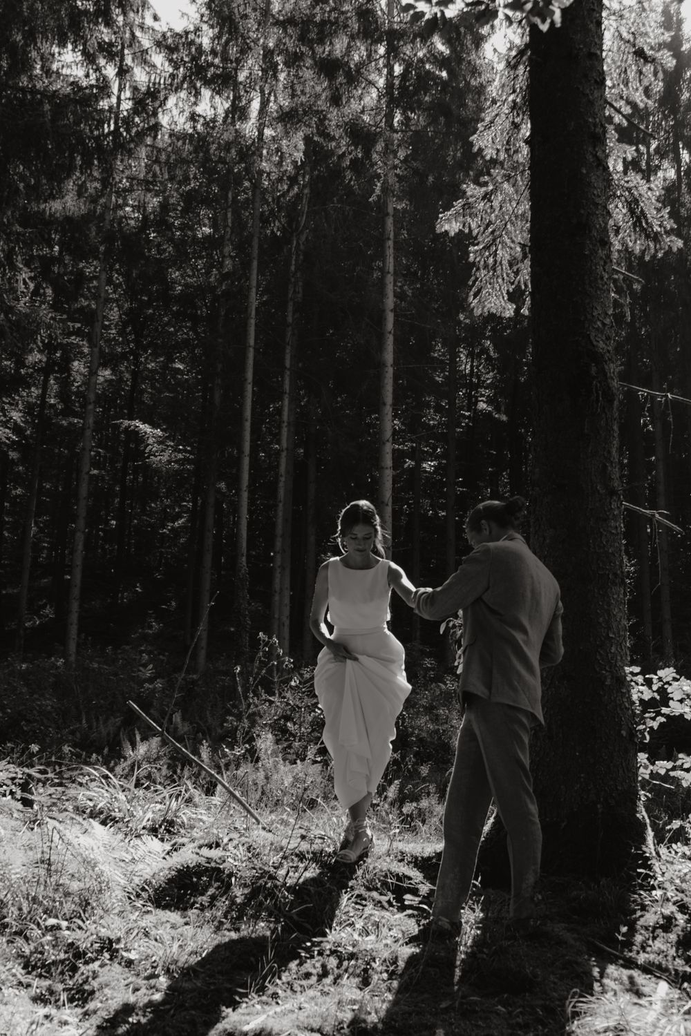 A couple in wedding attire walks through a sunlit forest.