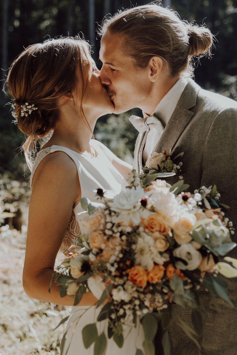 A couple kisses during their forest wedding ceremony while holding a peach and white floral bouquet.