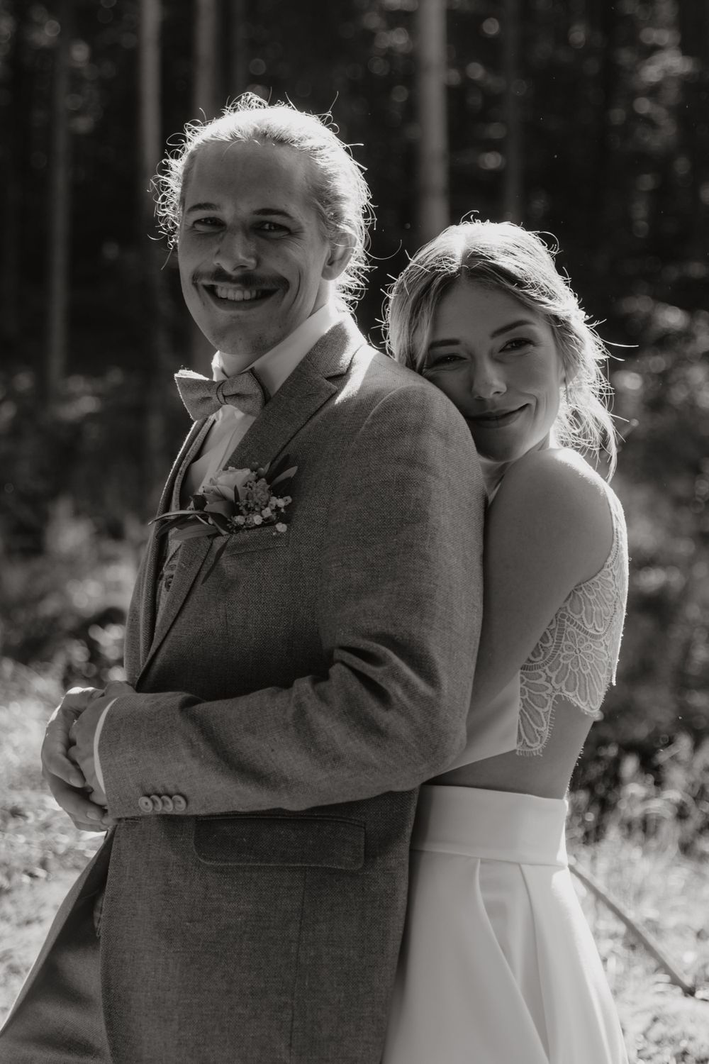 A bride and groom embrace while posing for a black and white portrait in a forest setting.