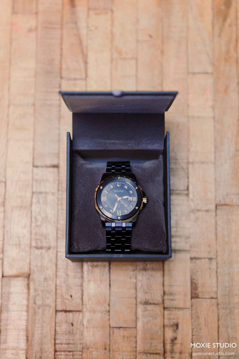 Black wristwatch displayed in open presentation box on wooden surface with studio lighting.