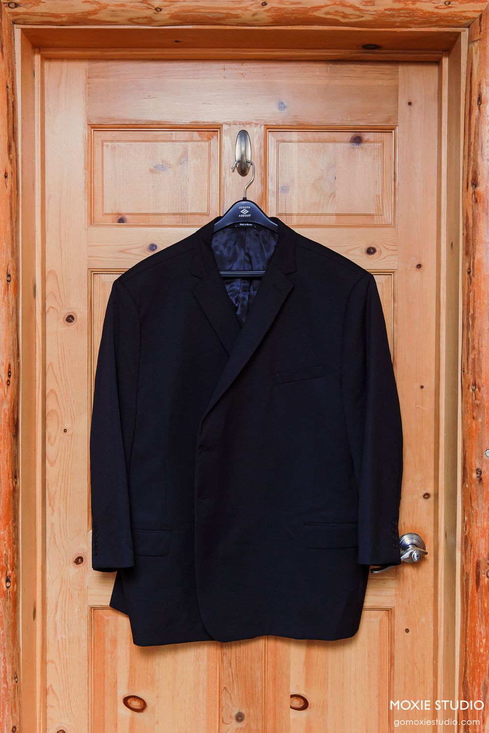 Black formal suit jackets hanging in a row on wooden hangers against a natural pine wood wall.