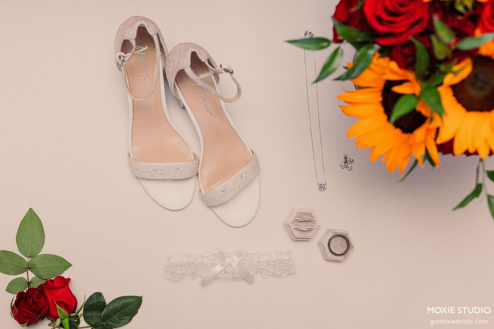 Wedding details including rings, sunflowers, red roses and elegant bridal shoes arranged on a light background.