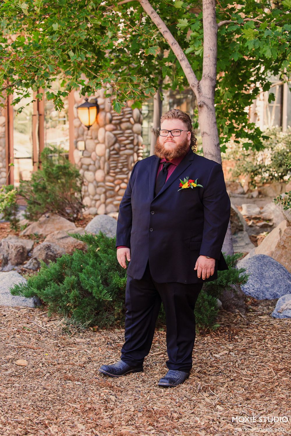 Person in formal attire poses in a garden setting with stone walls and trees in the background.