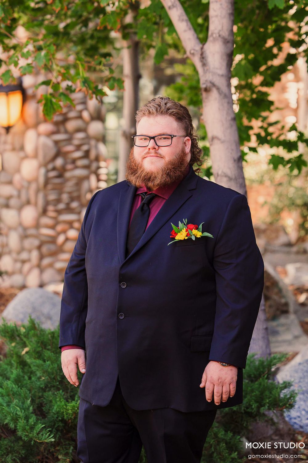 A groom in a navy suit with burgundy tie stands outdoors with a colorful boutonniere against a stone wall and trees.