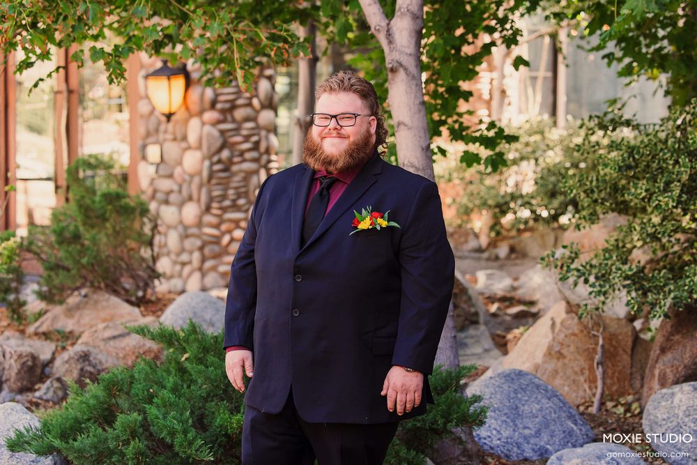 Person in dark suit stands among trees and landscaping with stone architecture visible behind them.