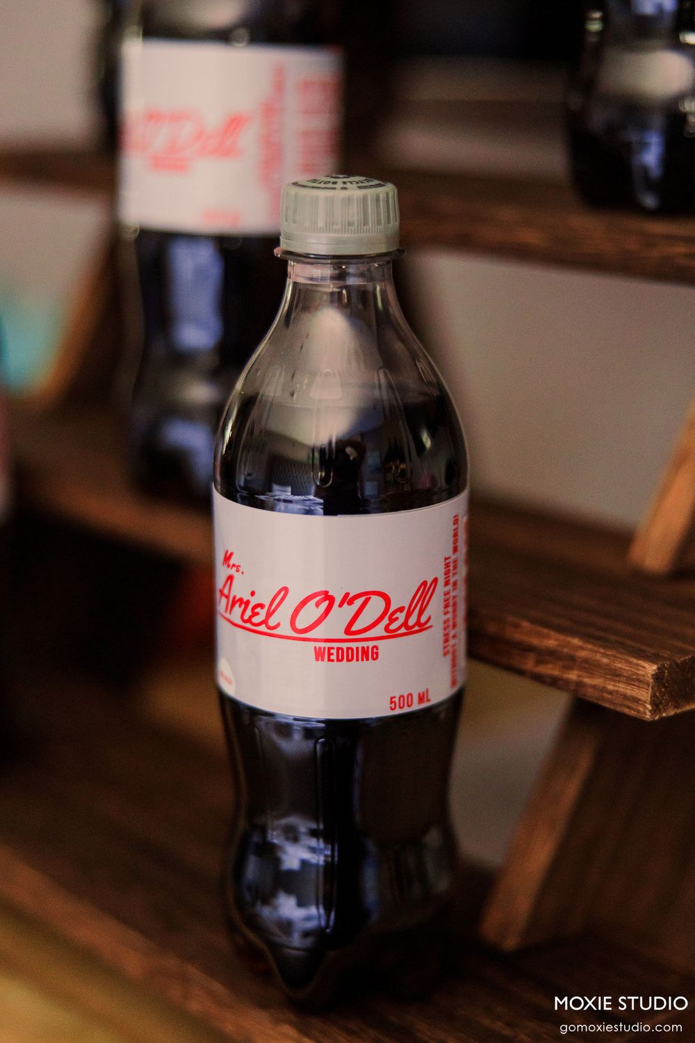 Row of personalized Coca-Cola bottles with custom wedding labels displayed on wooden surface.