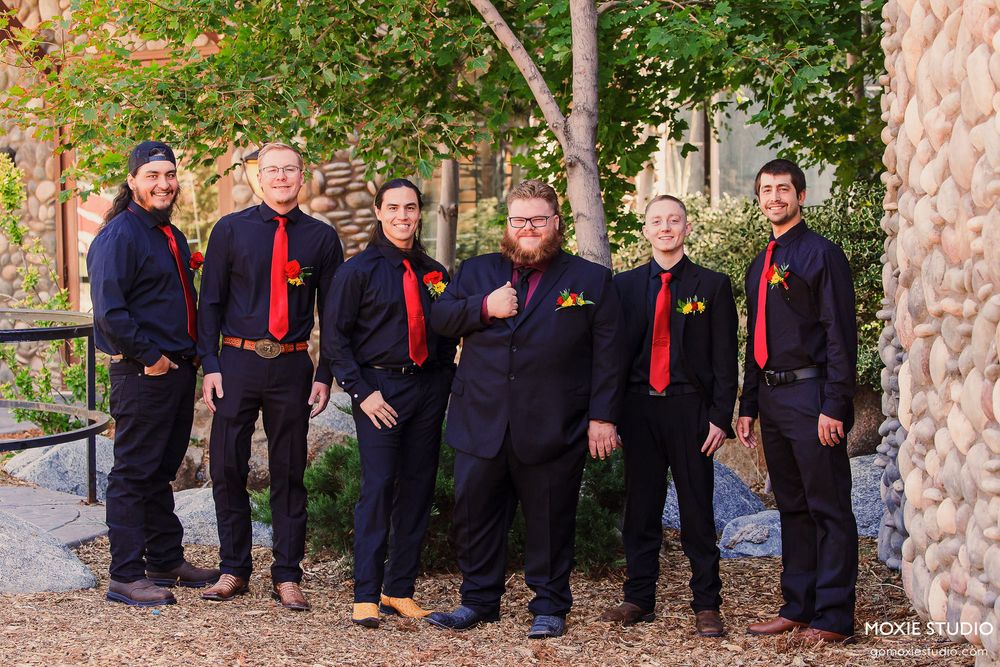 Wedding party members in black suits with red accents posing together outdoors near stone wall.