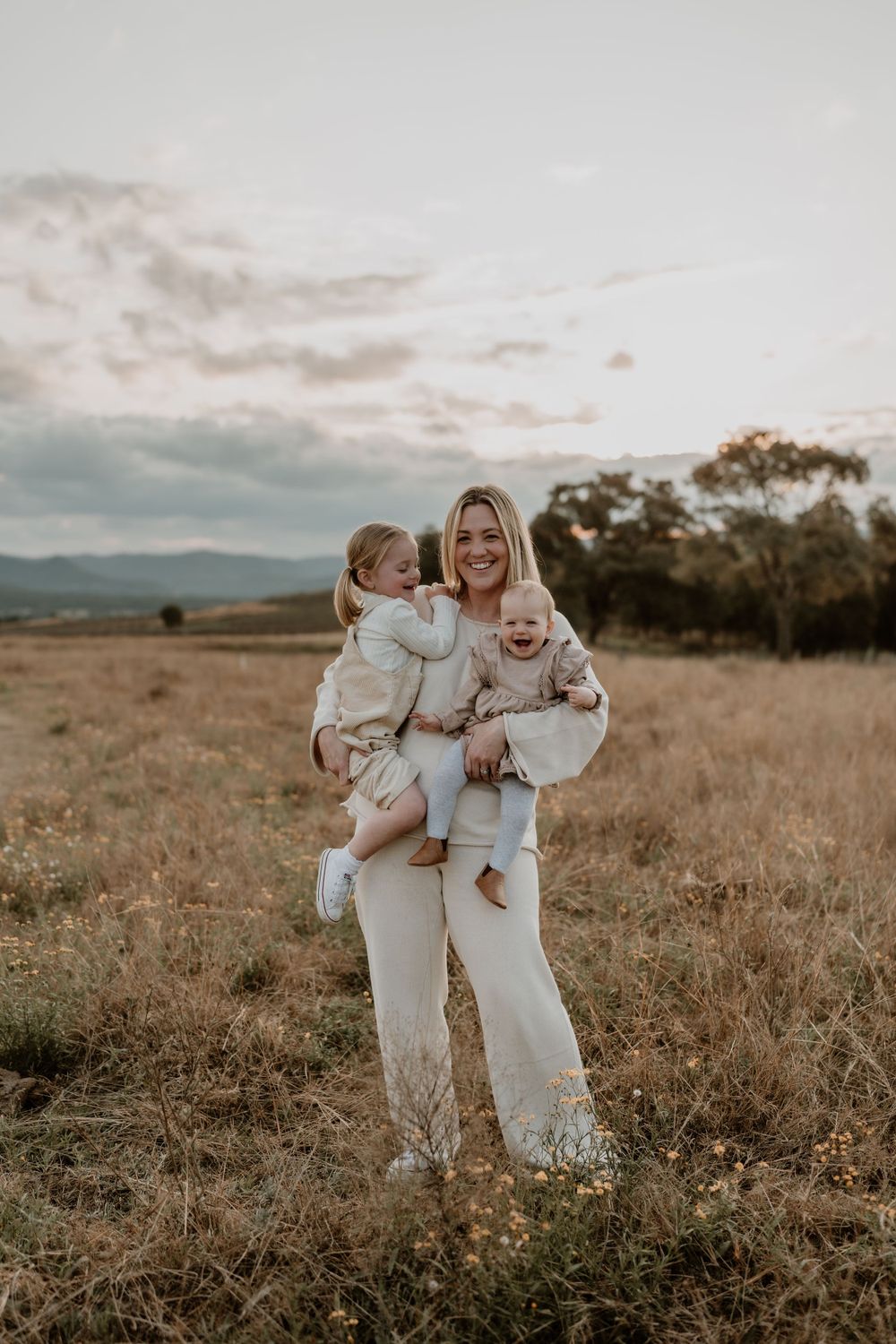 A smiling blonde mother holds her two young children in a sunlit open field.