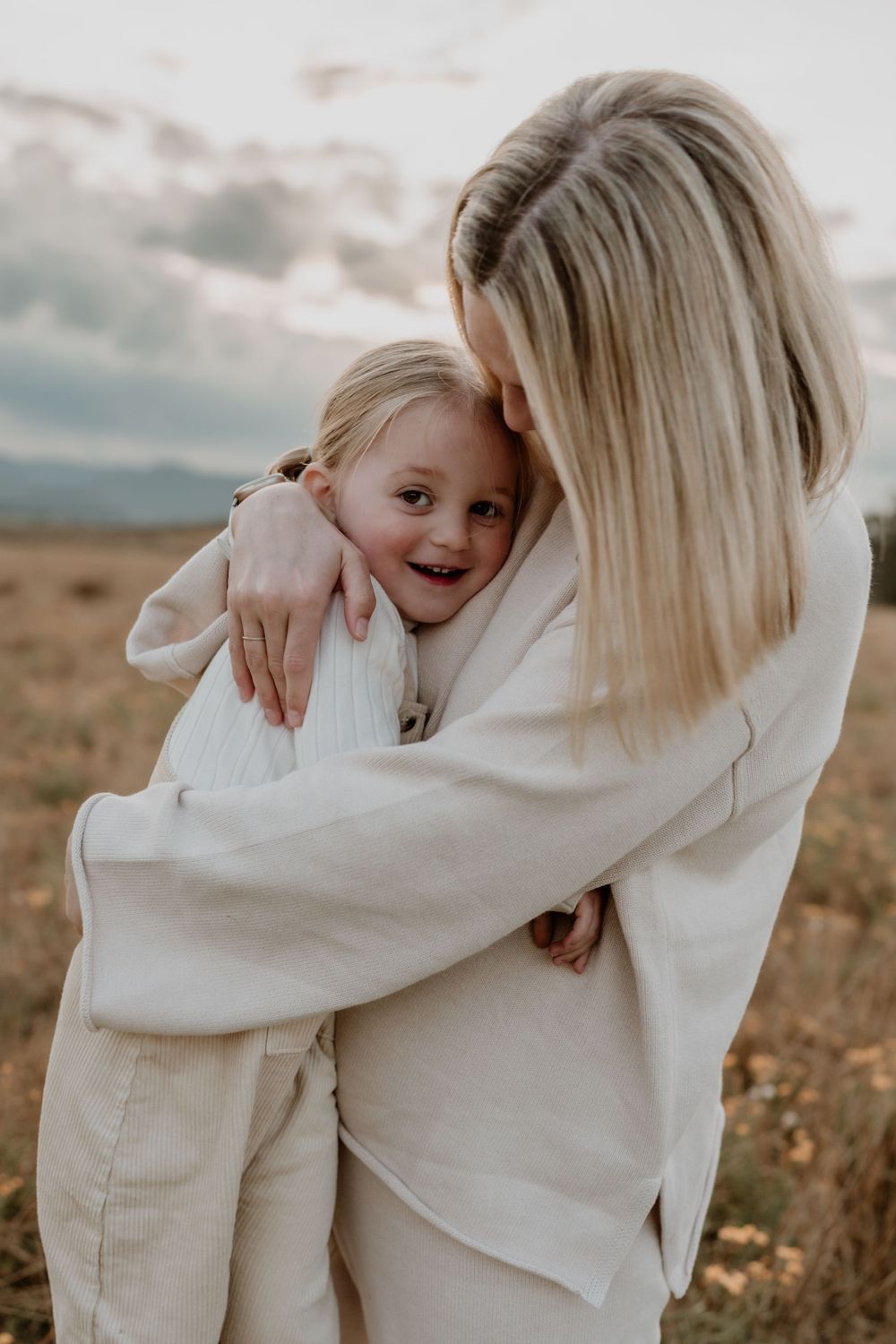 A mother wraps her arms around her laughing young daughter in an open field.