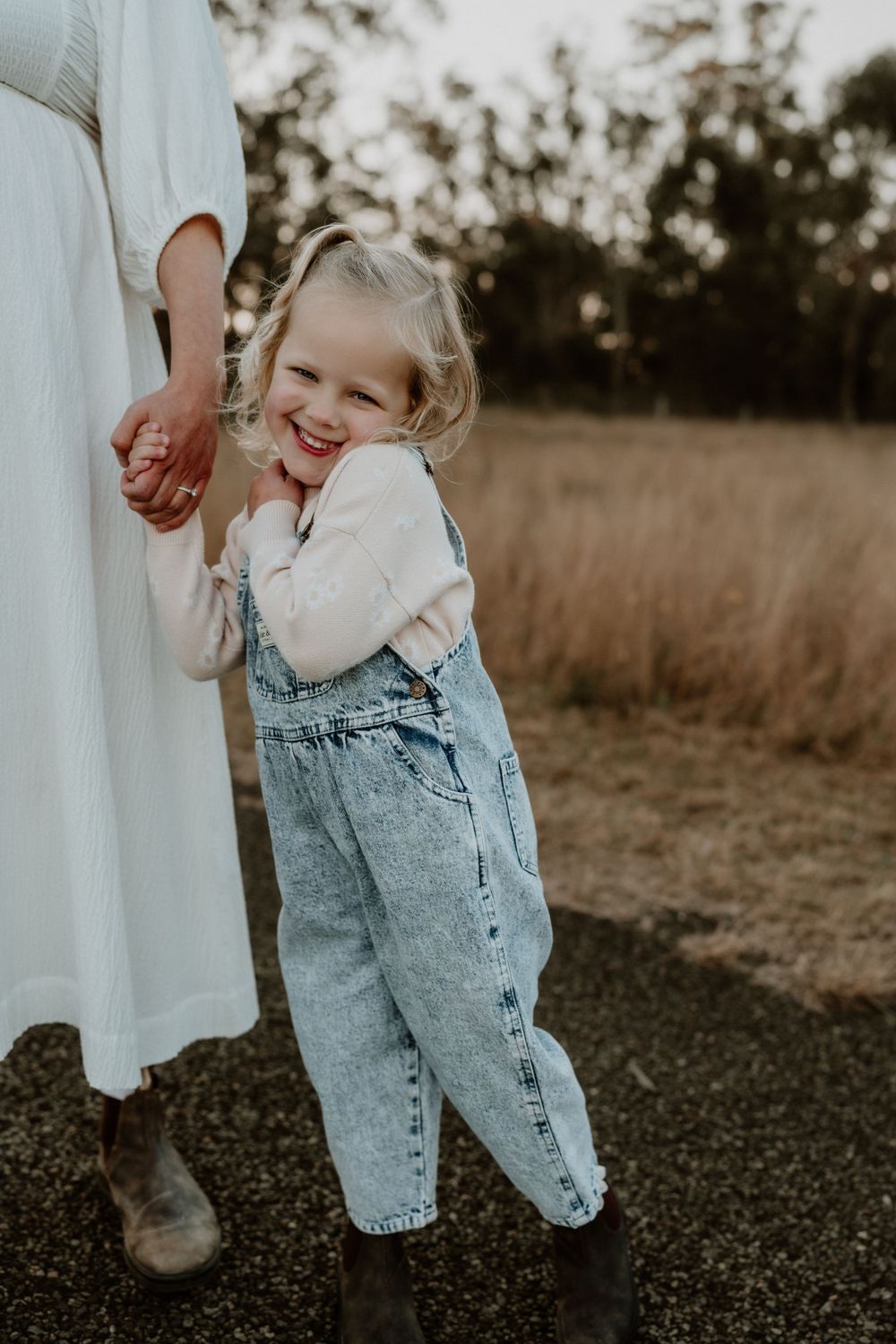 Happy toddler girl in denim overalls smiles while holding mothers hand outdoors.