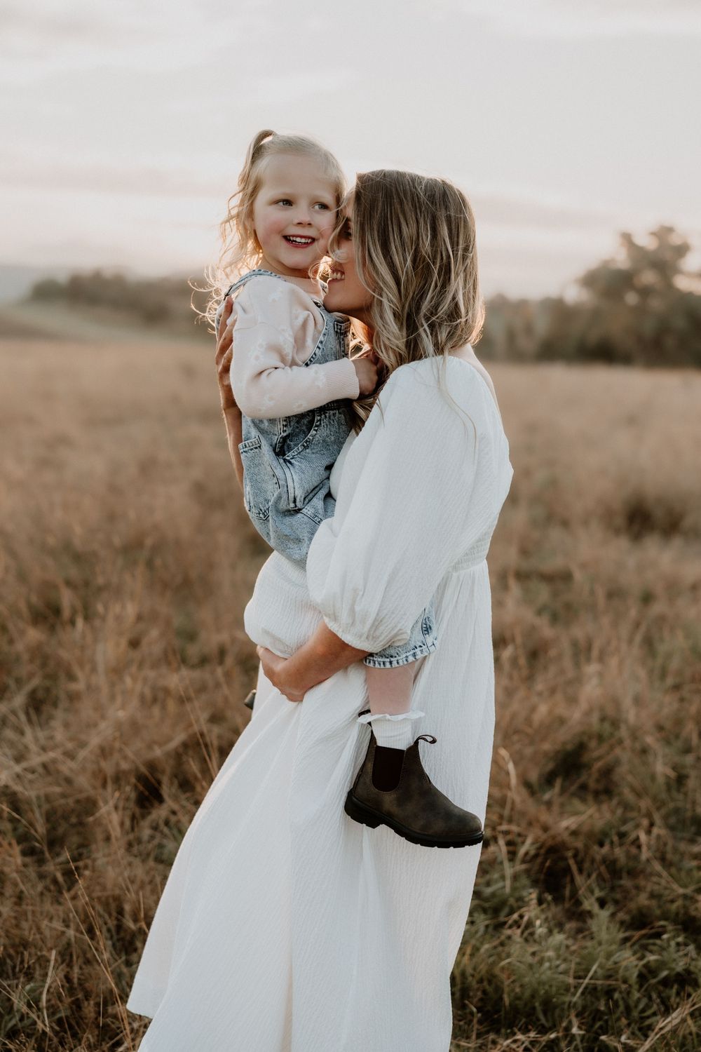 Mother in white dress kisses laughing little girl in denim overalls at sunset.