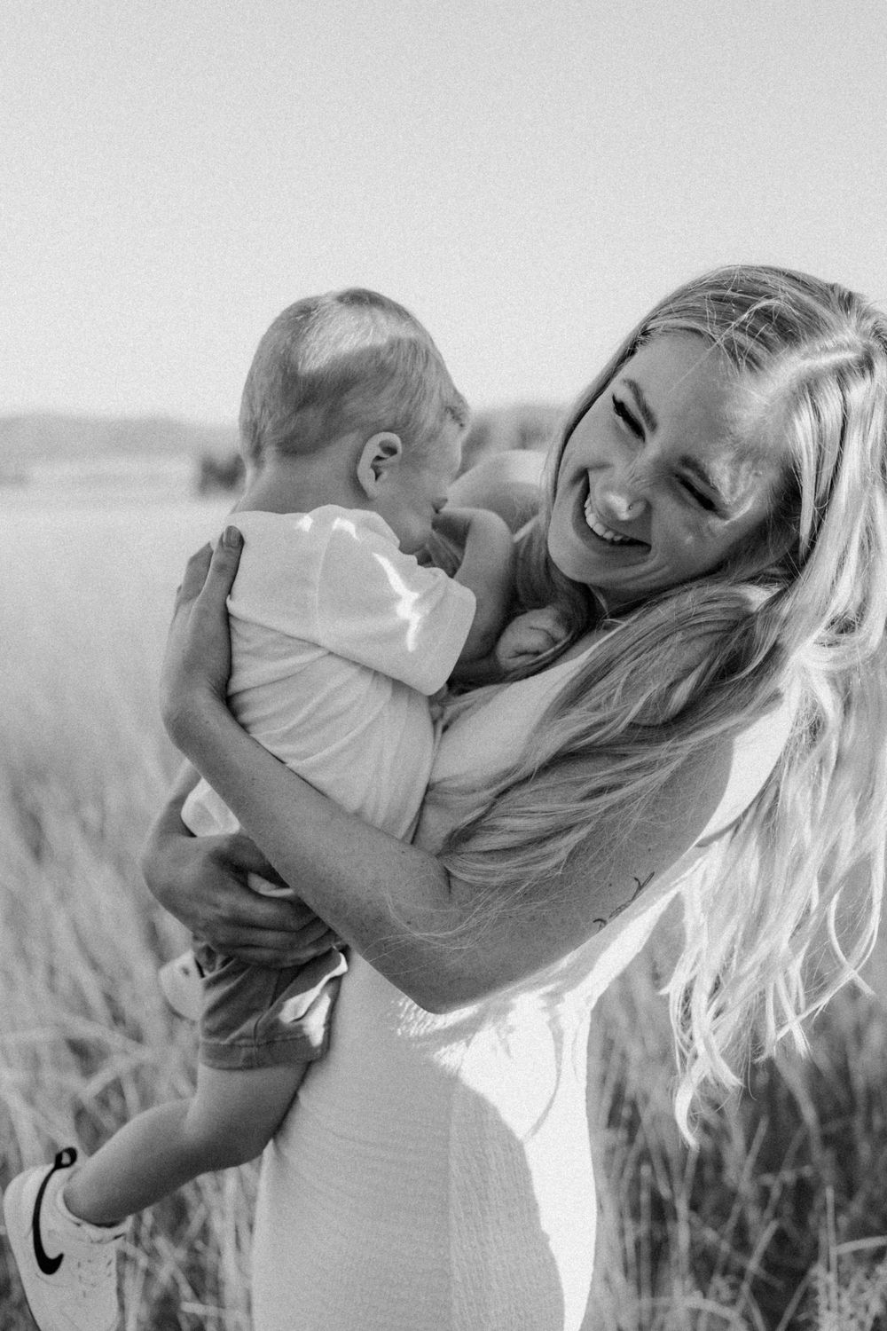 Black and white photo of joyful mother laughing while cradling newborn in field.