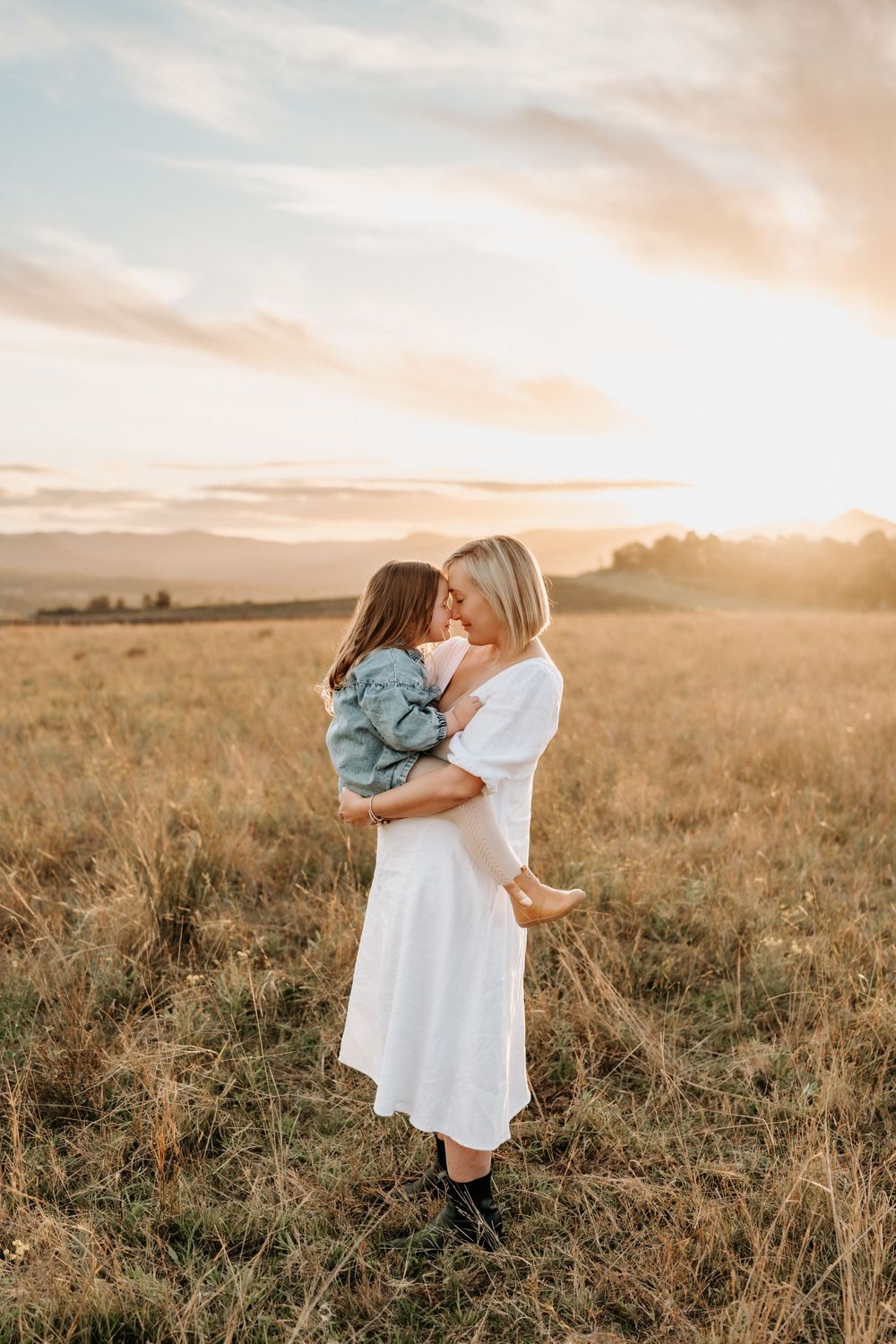 A mother in a white dress holds her young daughter in a golden field at sunset.