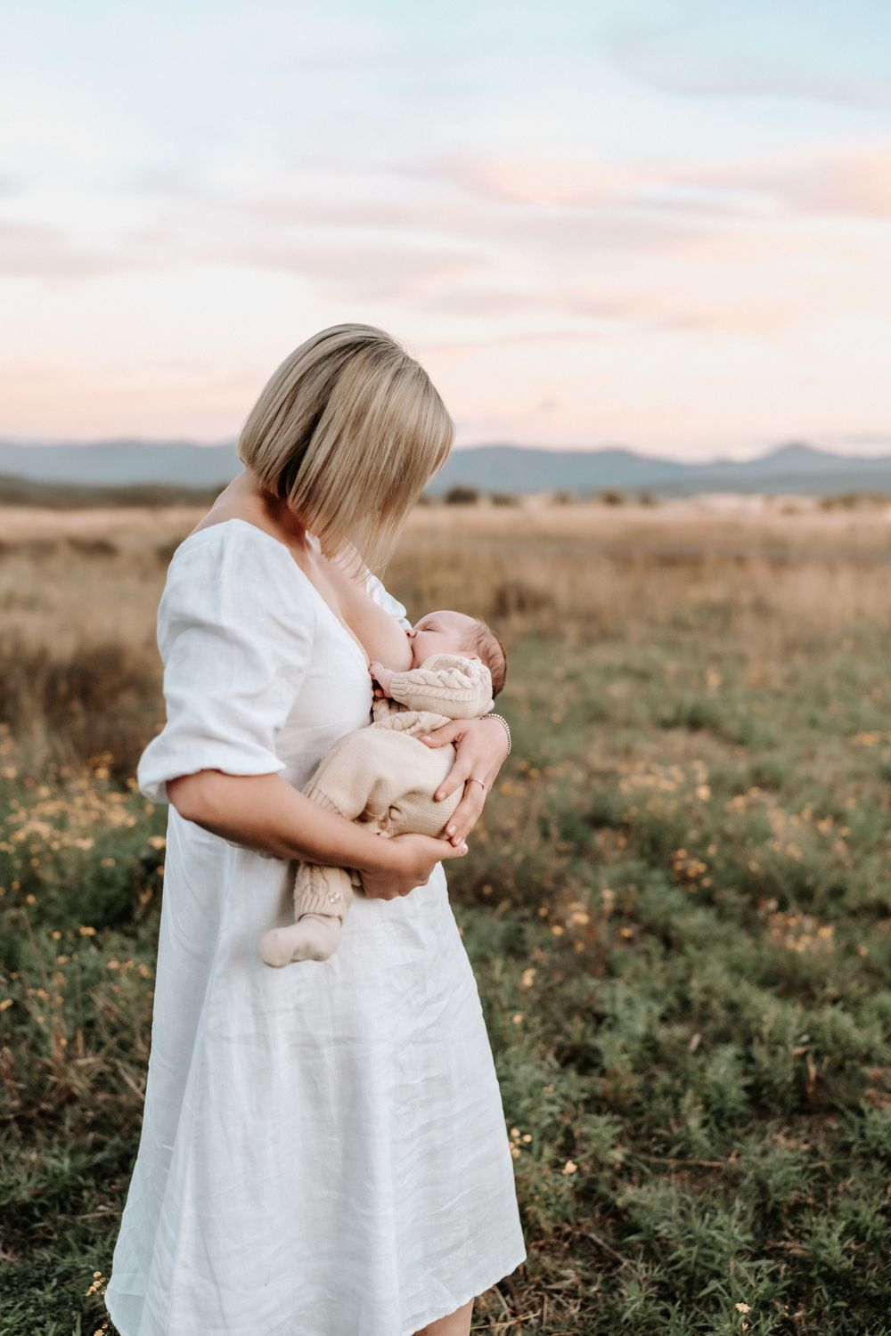A blonde woman in a white dress breastfeeds her newborn baby in a sunlit field.