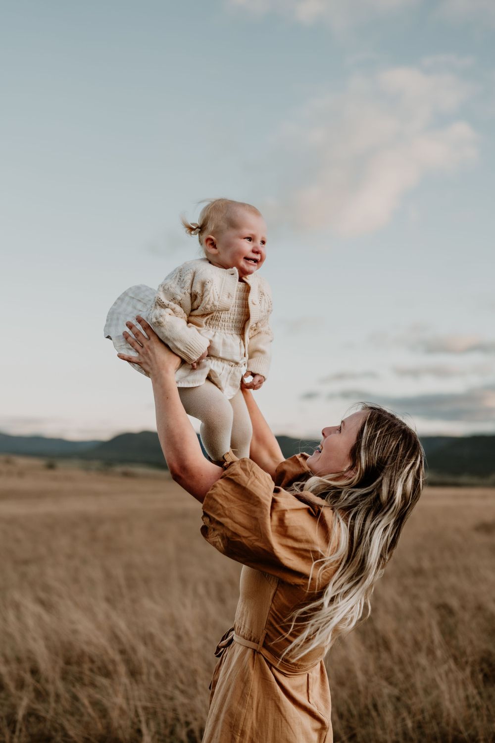Mother lifting smiling baby high in the air in golden field with mountains in background.
