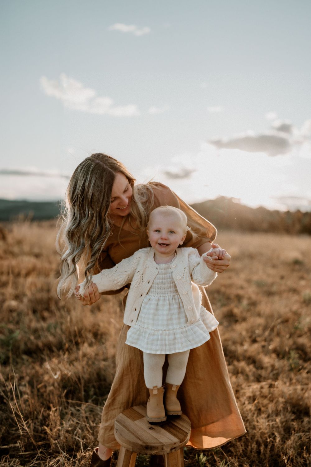 Mother in brown dress helping smiling toddler stand on stool in golden field at sunset.