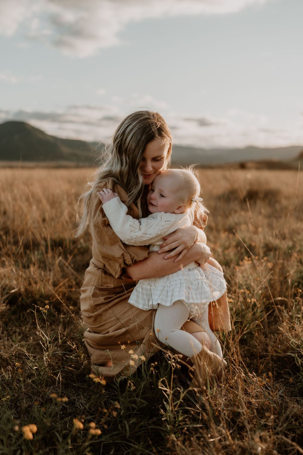Mother in brown dress hugging toddler daughter tightly in golden field at sunset.