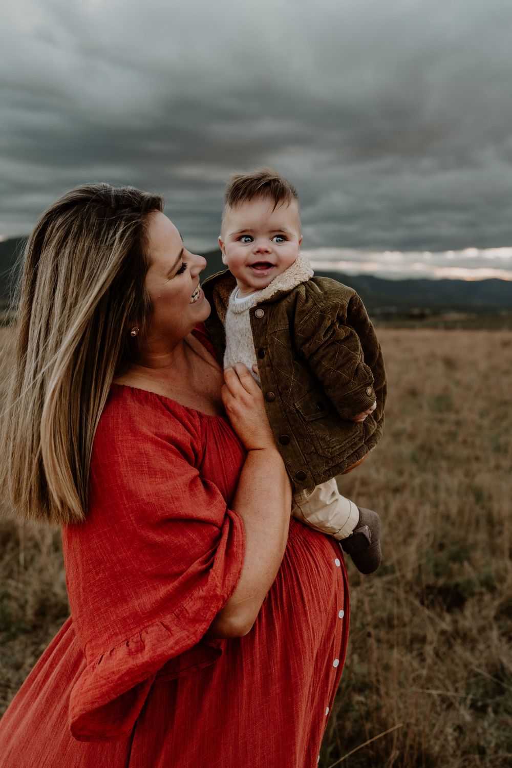 Mother in red dress holds smiling baby boy against dramatic cloudy sky outdoors.