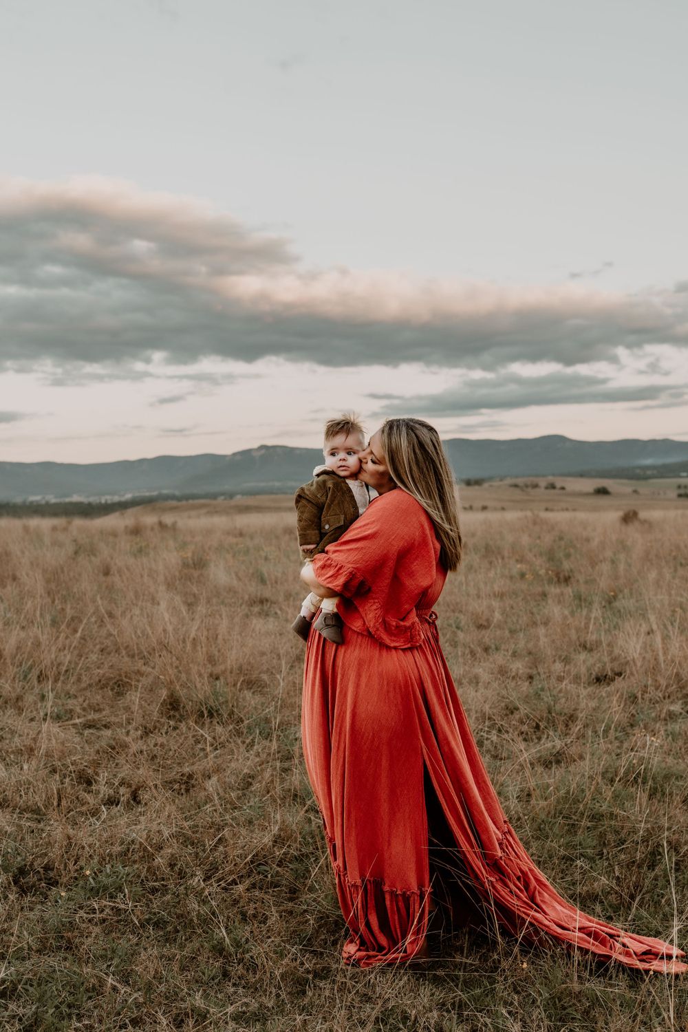 Woman in flowing red dress kisses baby while standing in golden mountain meadow.