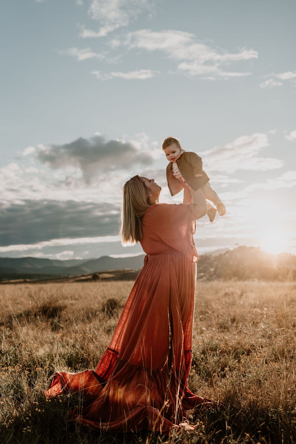 Mother in red dress lifts laughing baby toward golden sunset sky in open field.