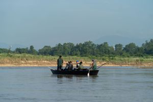 A small boat carries passengers across a wide river with green shoreline in the background.