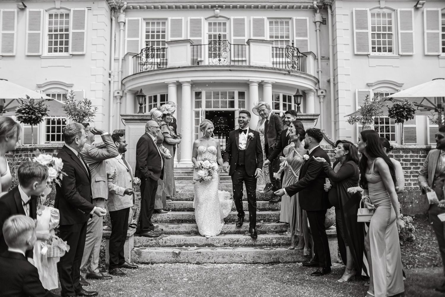 Wedding party and guests gathered in front of an elegant white colonial-style building.