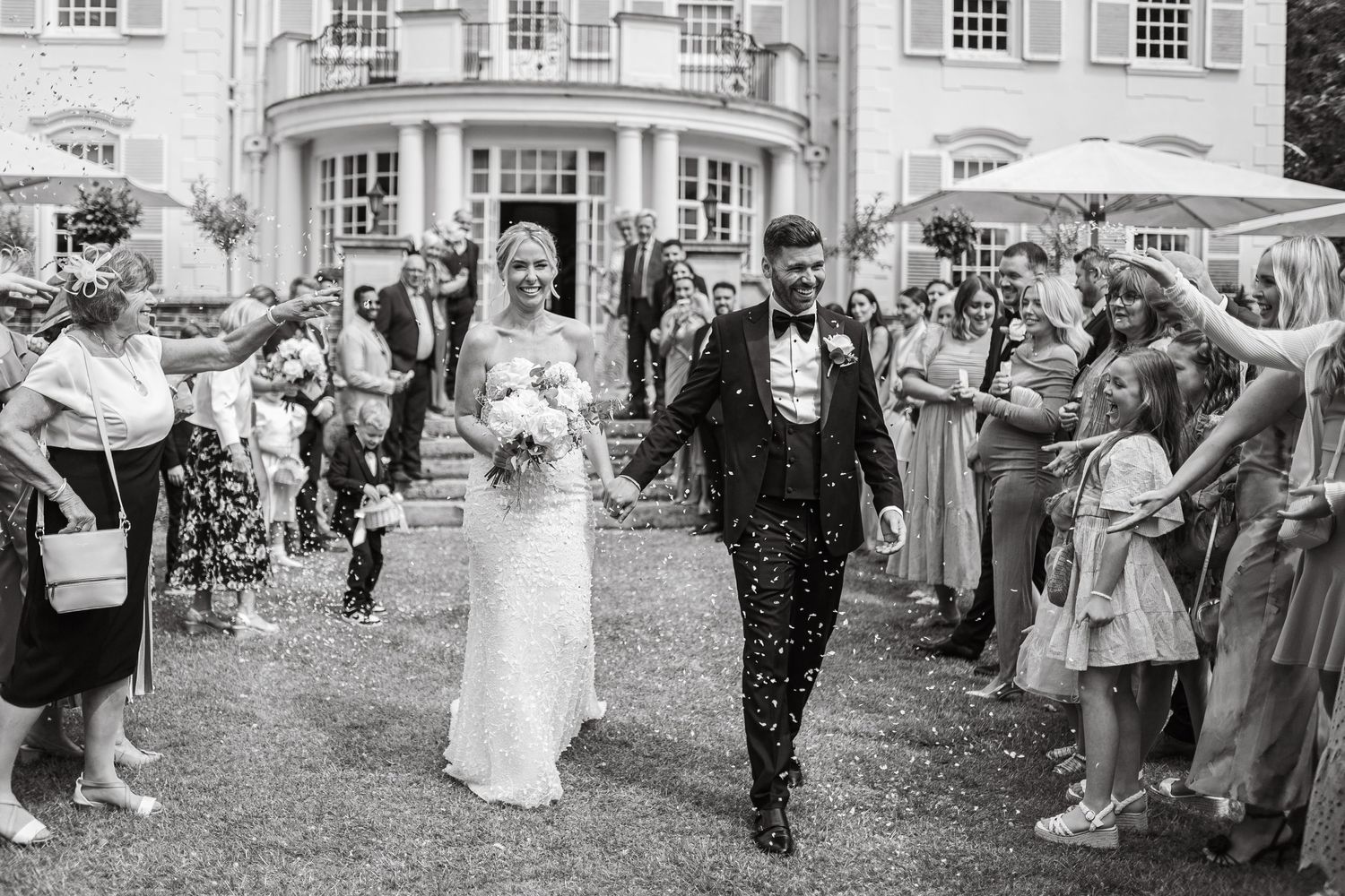 Wedding guests toss confetti at the newlyweds outside a stately mansion.
