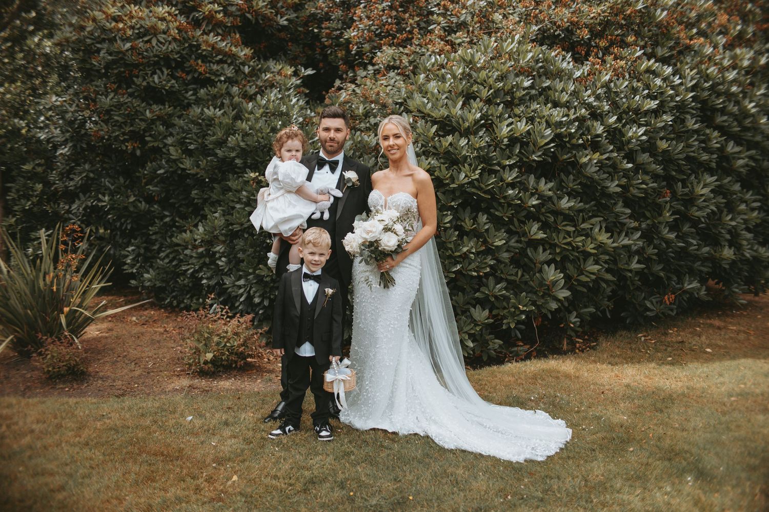 Wedding party poses for photos in front of lush garden hedges.