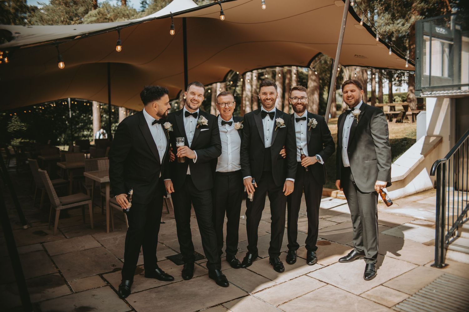 Groomsmen in black tuxedos pose together outdoors under string lights at a wedding venue.
