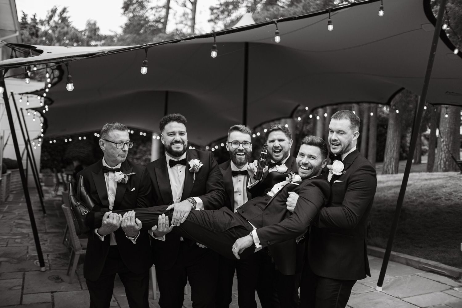 Wedding party members stand together on a patio with string lights overhead.