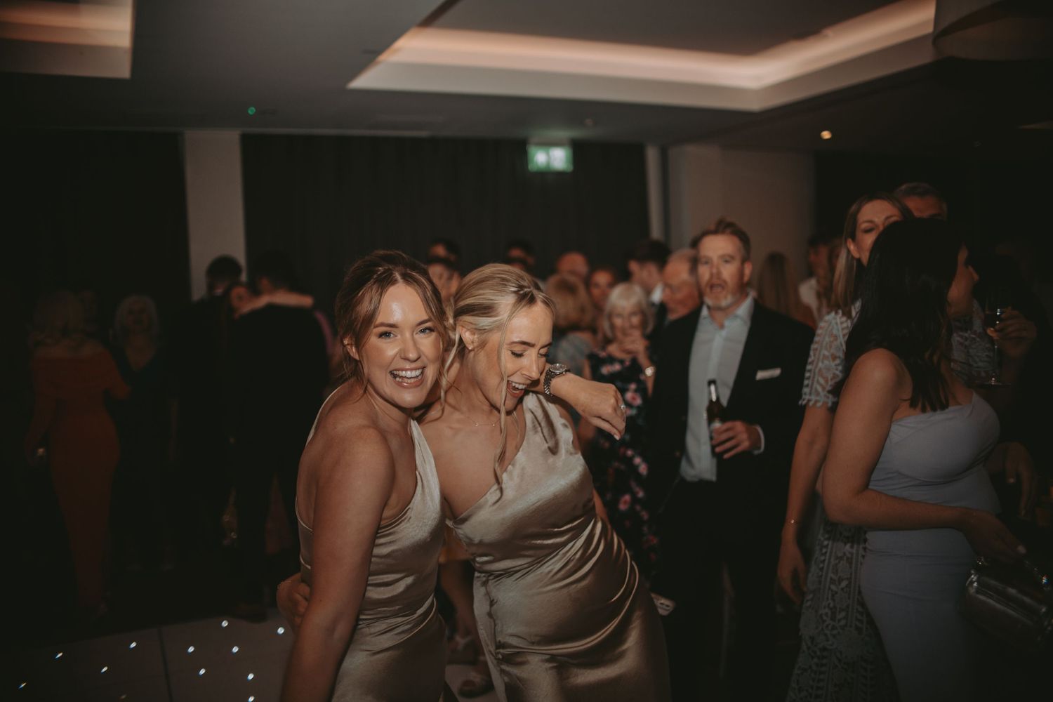 Wedding reception guests dance and celebrate in a dimly lit ballroom with ambient lighting.