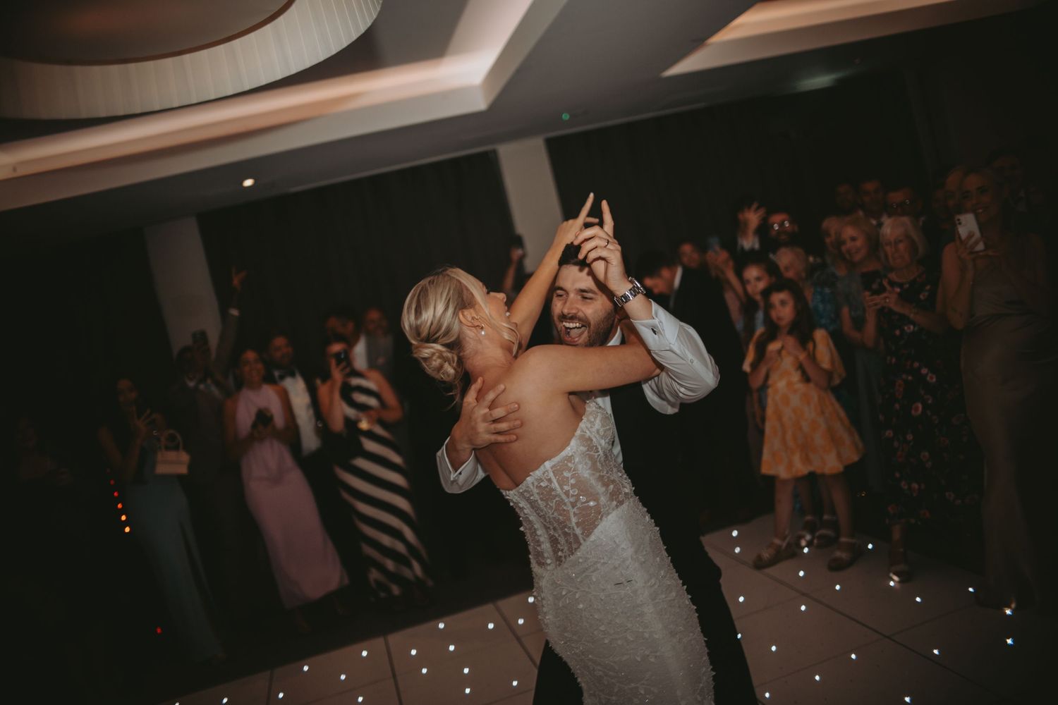 Wedding guests celebrate on a dance floor illuminated by overhead string lights.