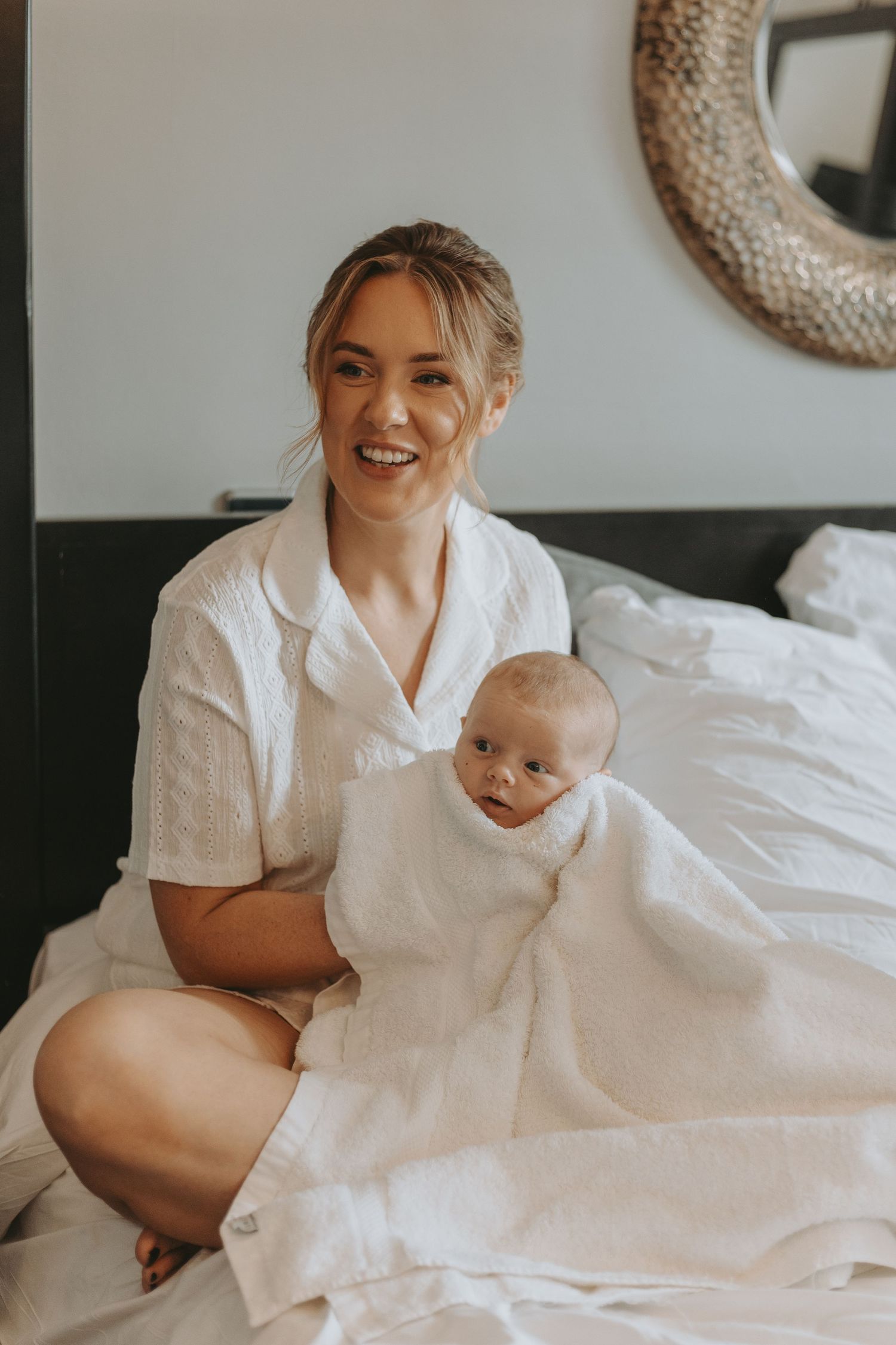 Mother and newborn baby dressed in white sitting together on a bed with a decorative mirror in the background.