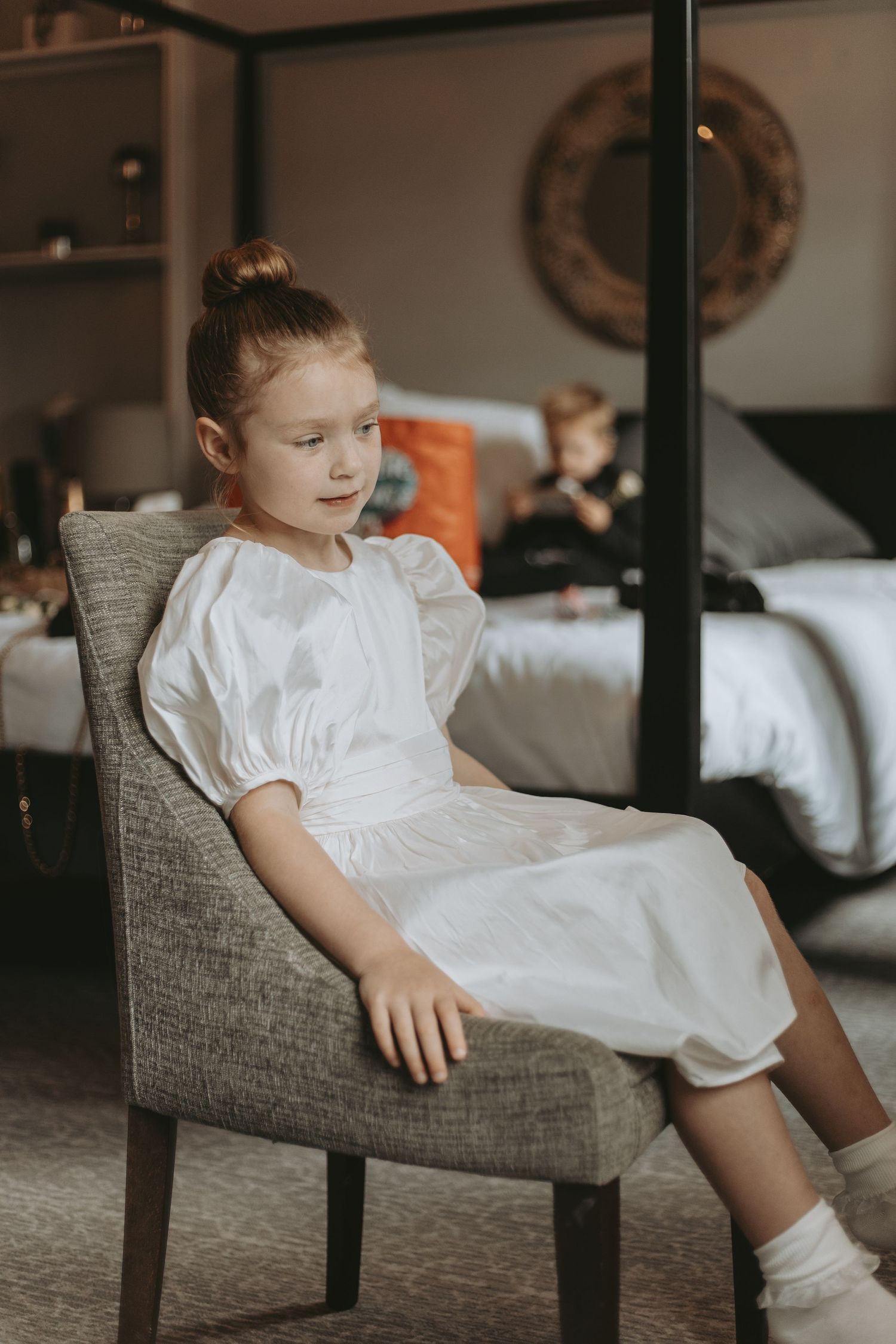 Young child in white dress sitting in gray armchair during wedding preparations.
