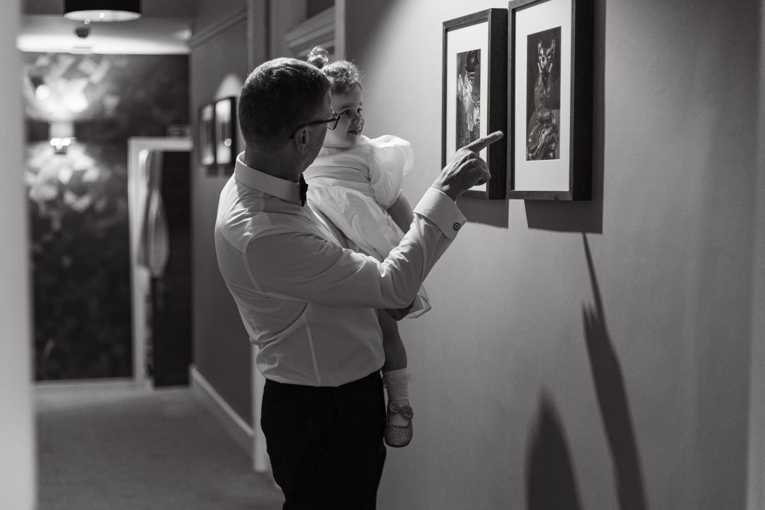 Person in formal attire holding child while looking at framed artwork in hallway.