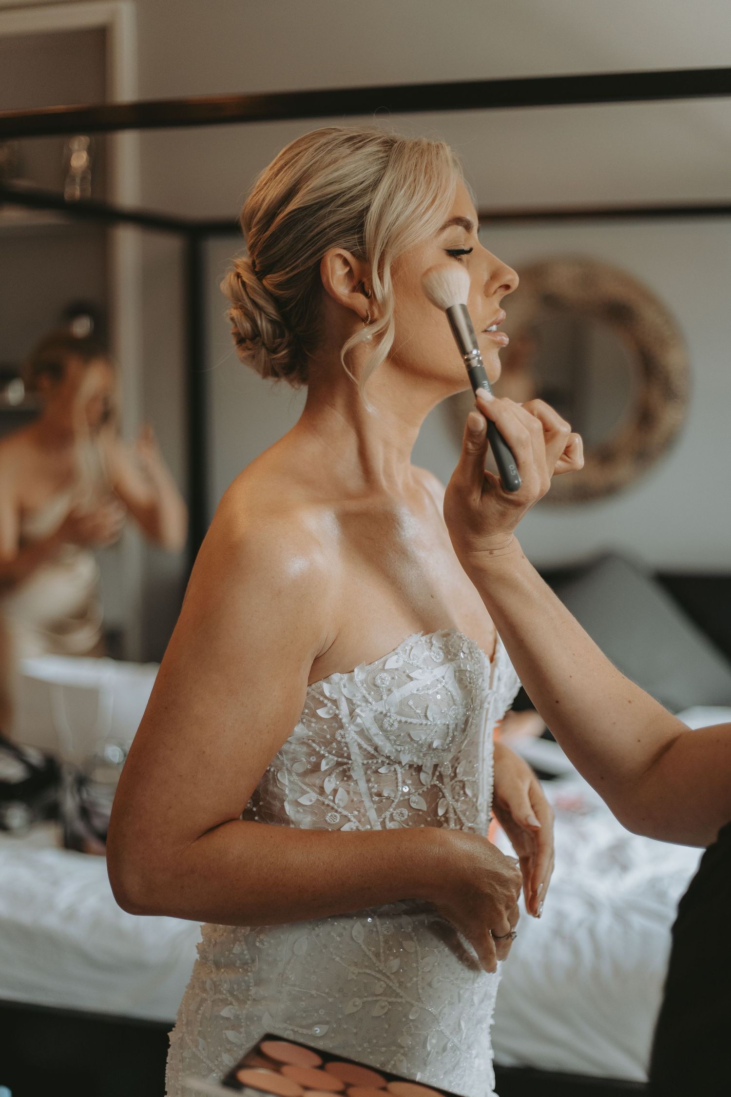Bride in lace wedding dress getting final makeup touches during preparation.