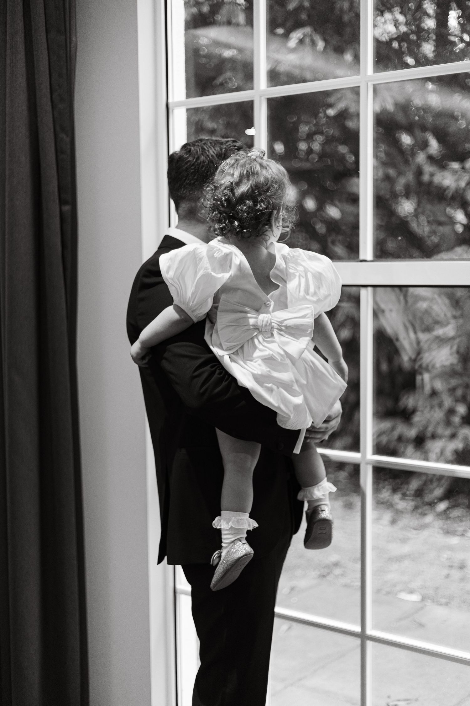Person in formal suit holding child in white dress by window.