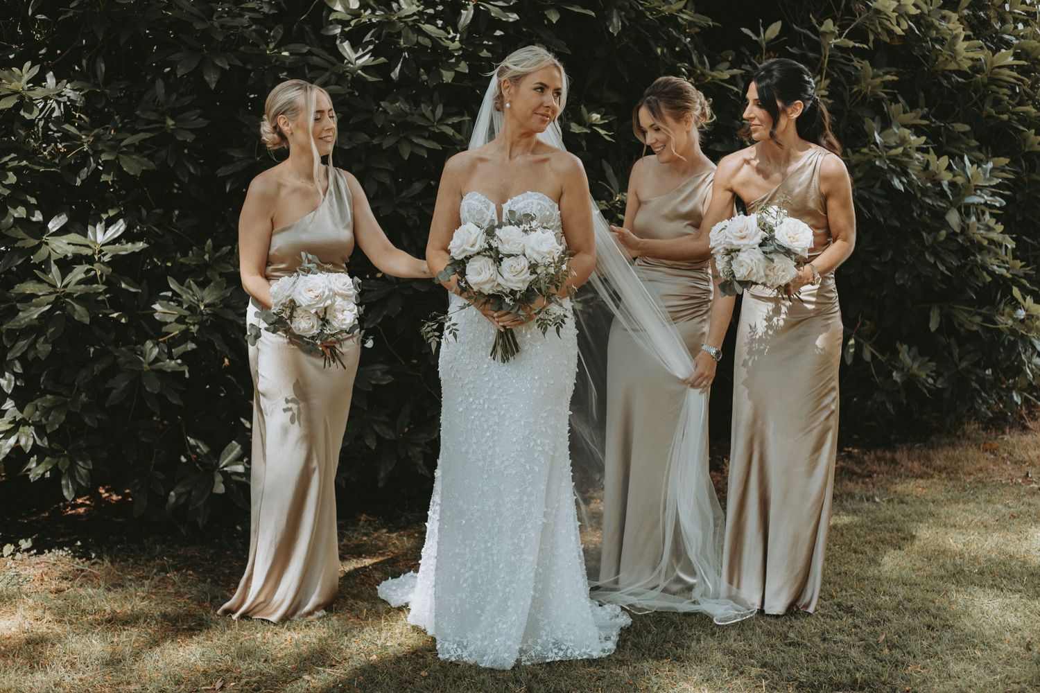 Wedding party members in neutral dresses gather outdoors holding white floral bouquets.
