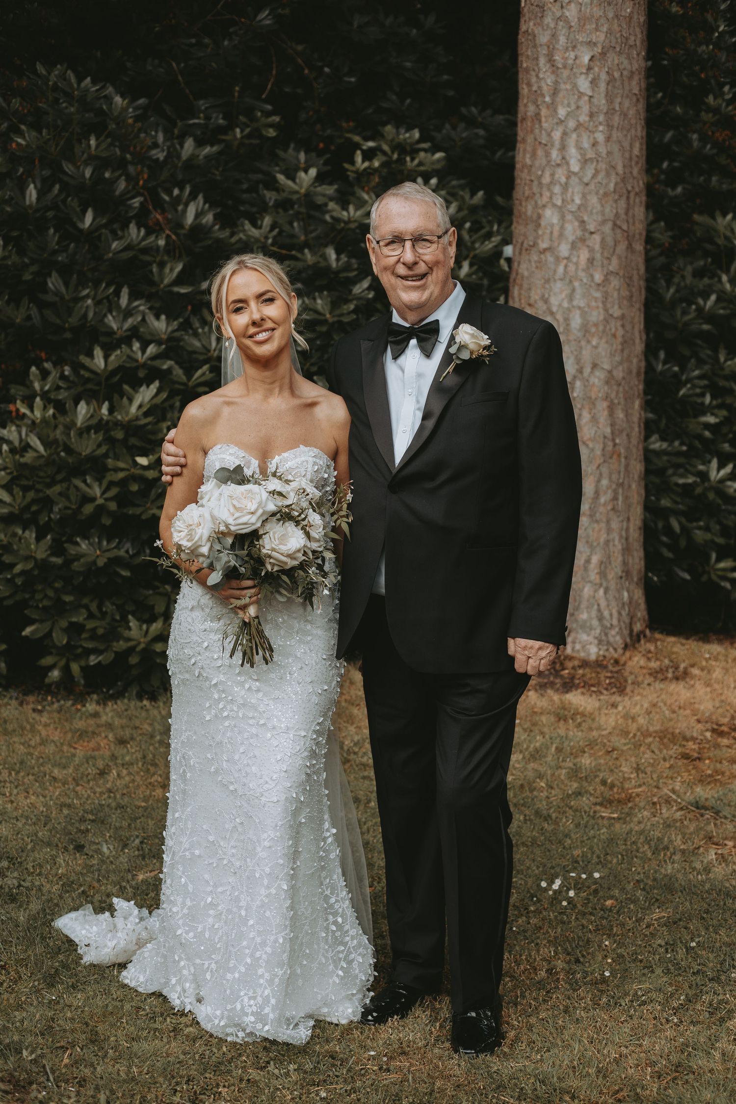 A bride in a sequined white gown poses with family member in formal attire under tall trees.