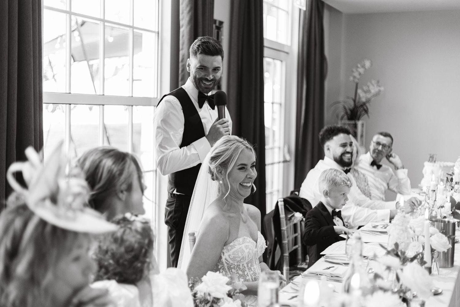 Wedding guests celebrate and laugh together during a joyous reception in a bright room with large windows.