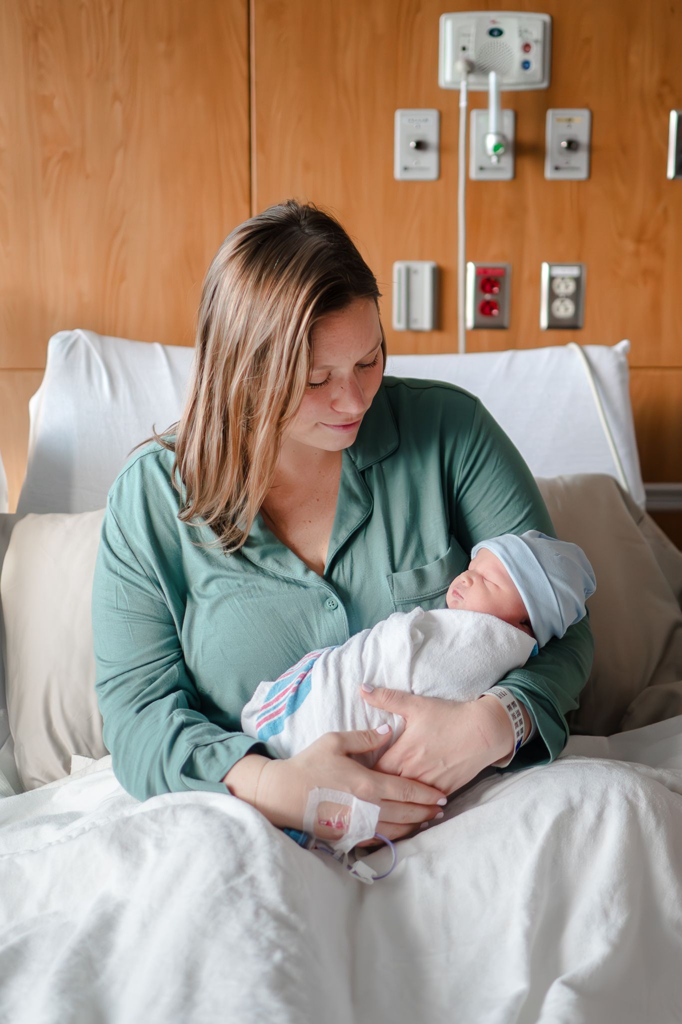 A woman wearing a teal blouse sits in a hospital bed, holding and gazing lovingly at a newborn baby swaddled in white.