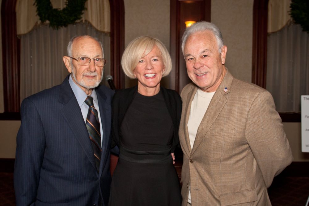 Three elegantly dressed people pose together at a formal event with holiday decorations.