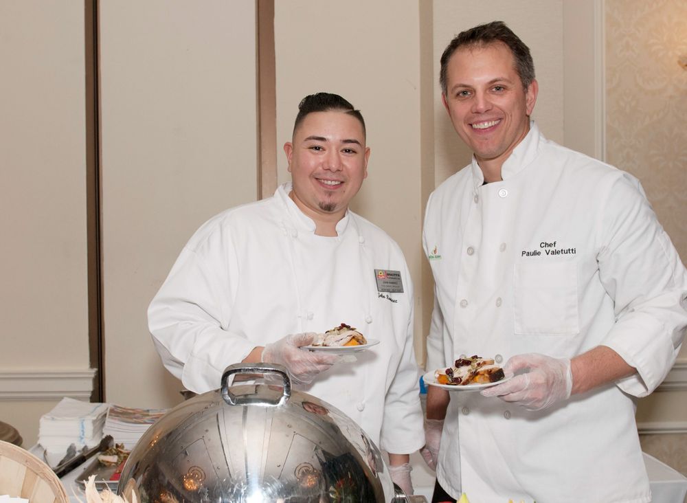 Two chefs in white uniforms posing with food service equipment at catered event.