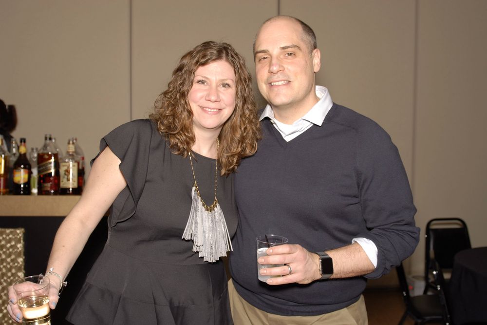 Two people pose together at an event with a bar counter in the background.