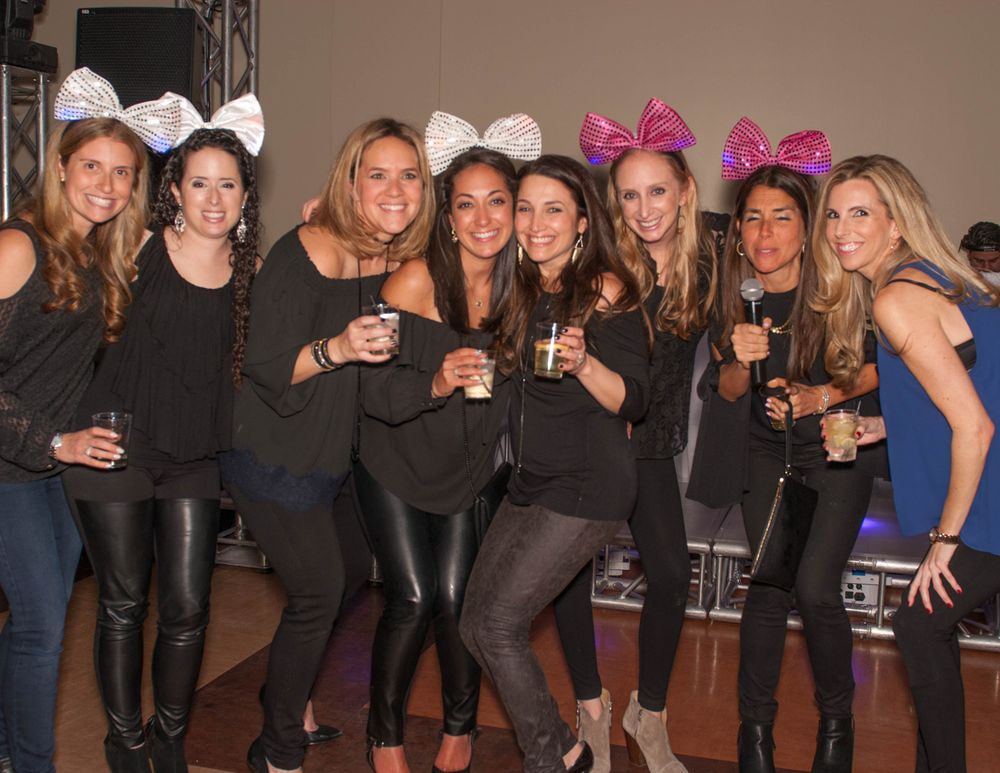 A group of friends in black outfits wearing decorative bows pose together while holding drinks at a party.