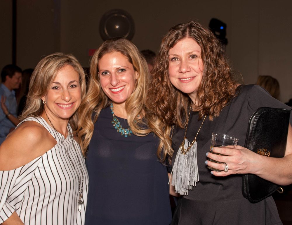 Three friends smile while holding drinks at an indoor social gathering.