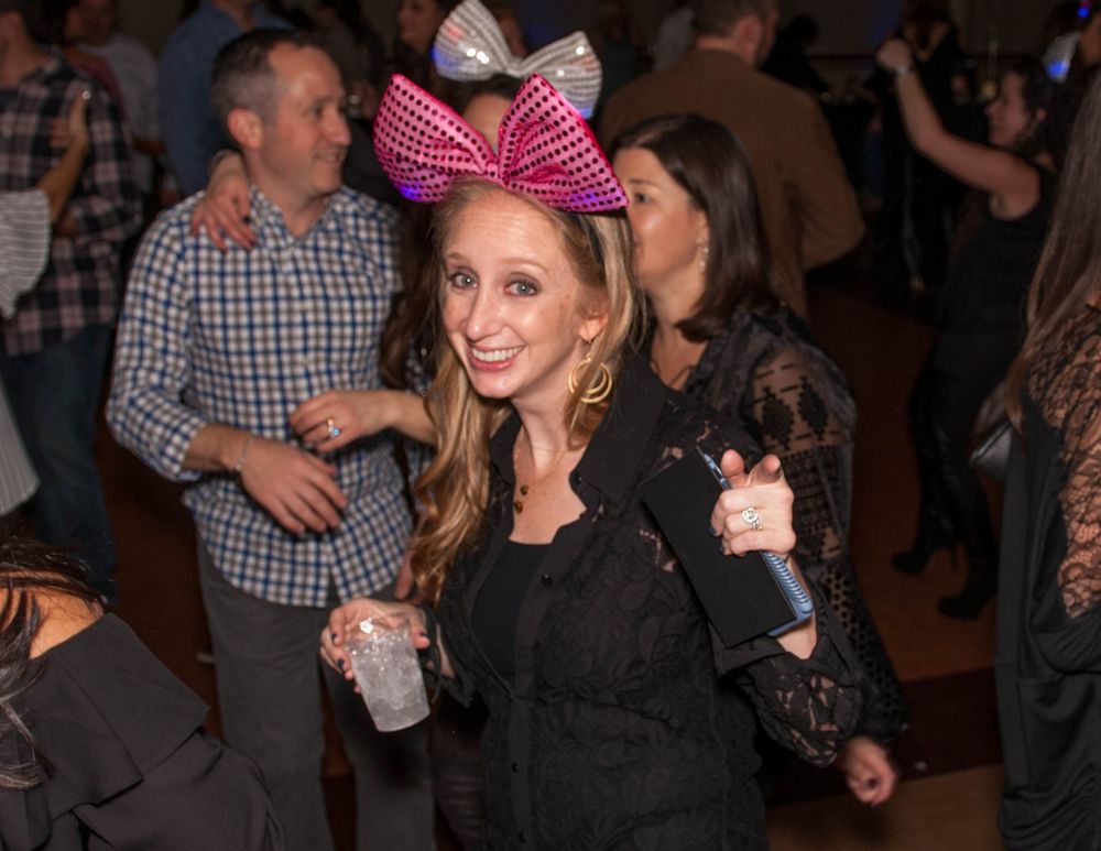 A person wearing a black outfit and large pink bow poses with a drink at a party.