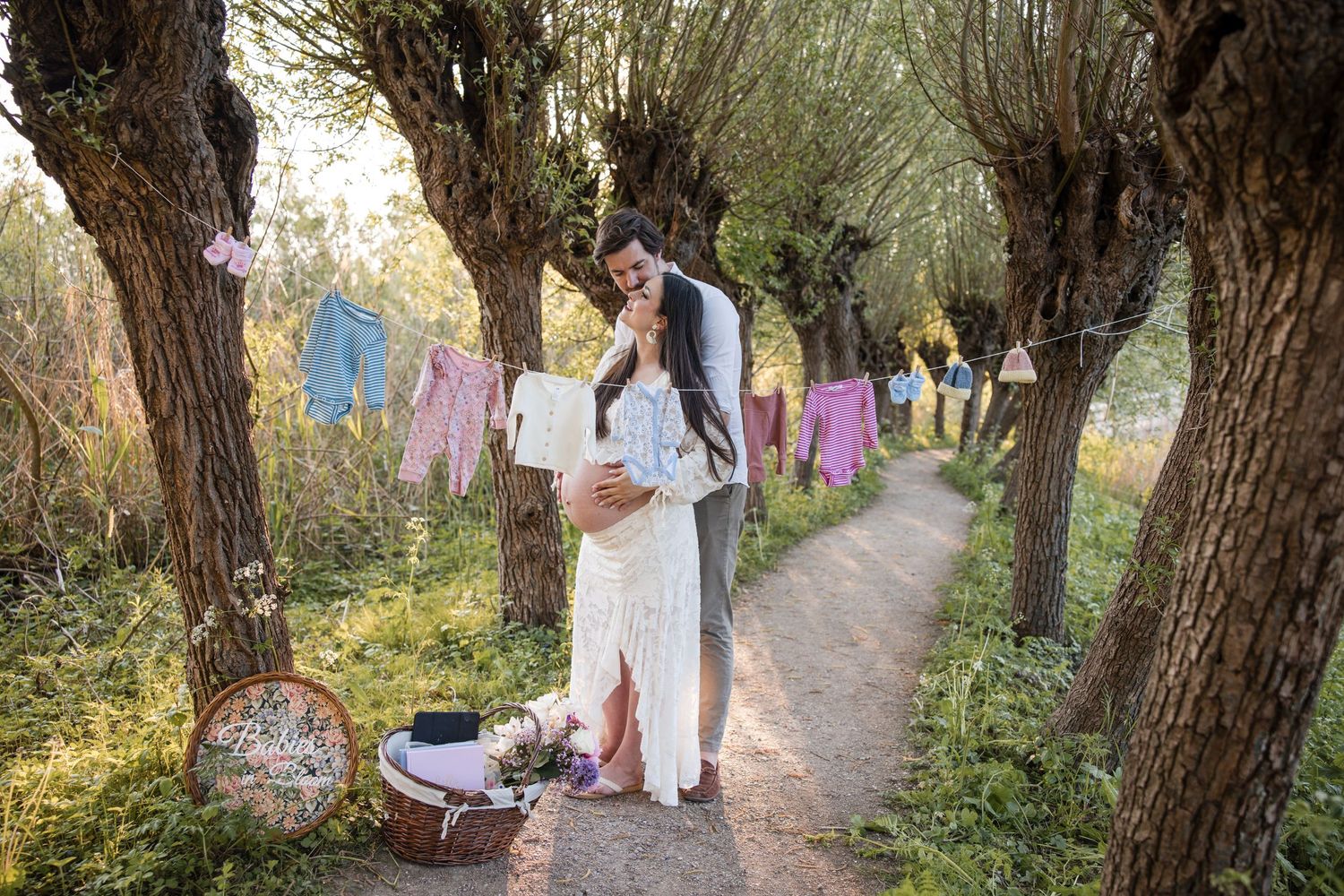 A pregnant couple stands on a dirt path near trees with baby clothes hanging on a clothesline and a basket of laundry nearby.