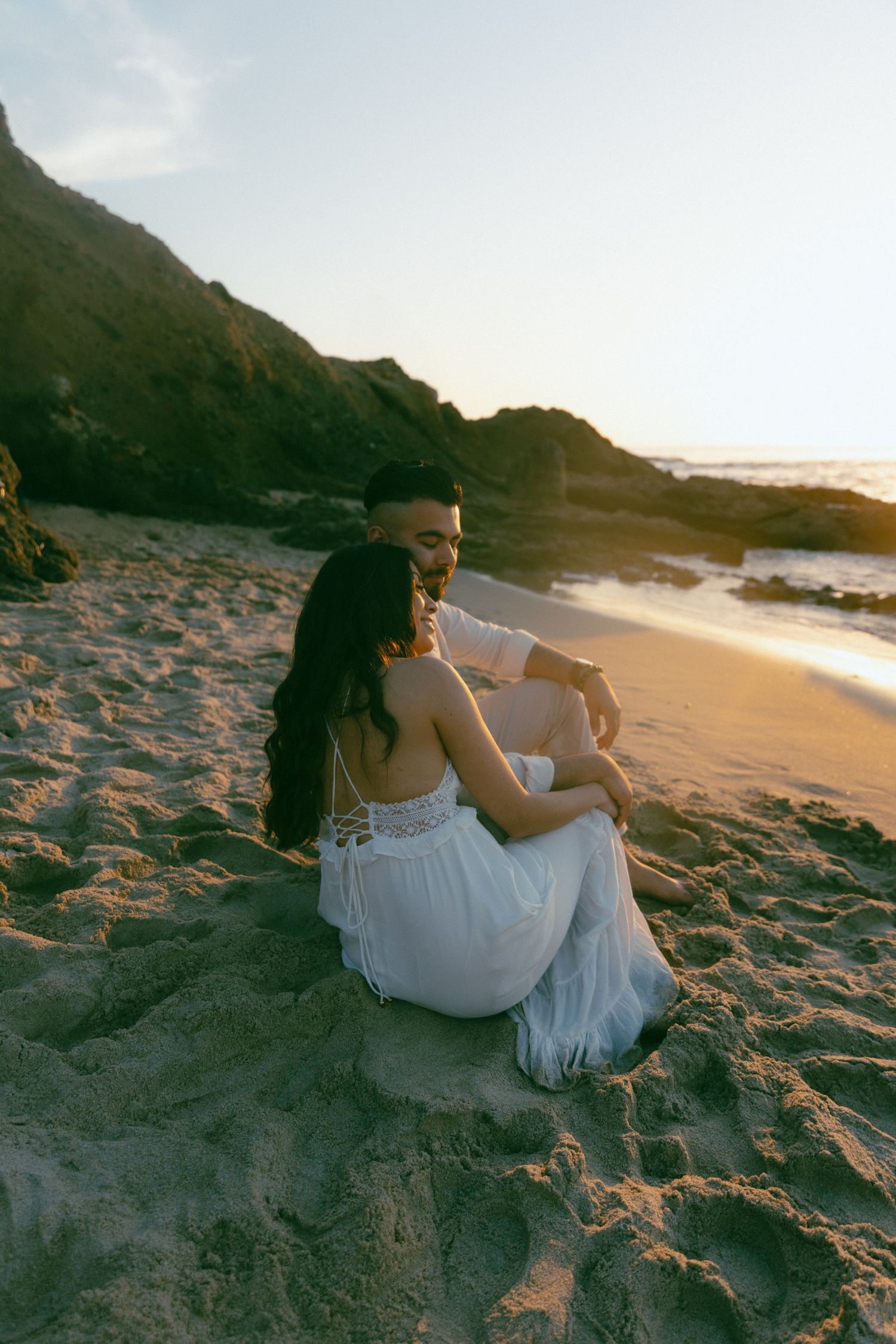 A couple sits close together on a sandy beach at sunset with rocks and ocean waves in the background.
