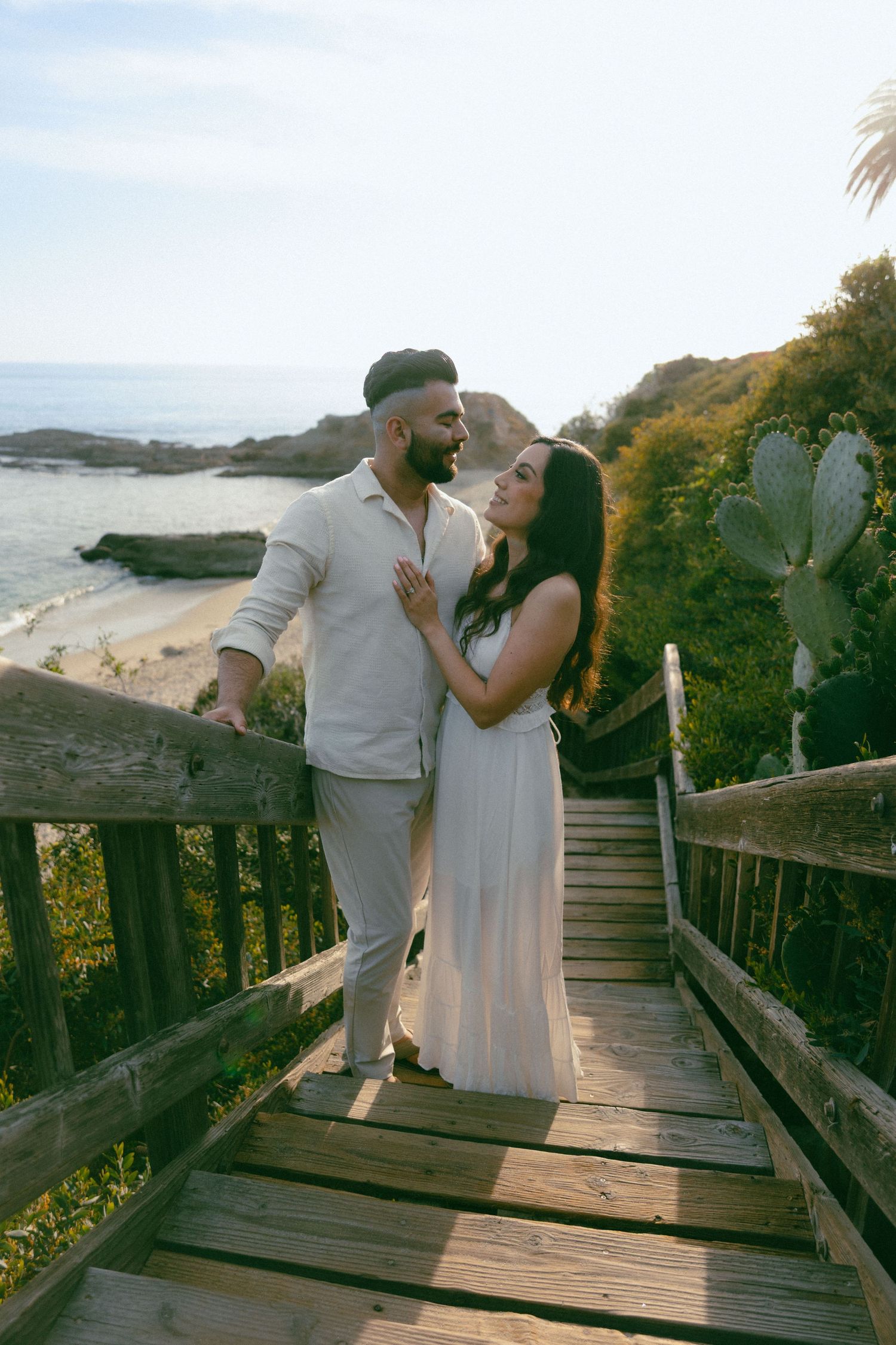 Romantic couple shares tender moment on wooden boardwalk overlooking scenic beach with cactus and ocean view at sunset.
